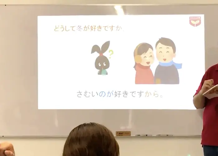 Salón de clases de japonés, con alumnos y maestro. Se ve una diapositiva de una imagen en japonés.